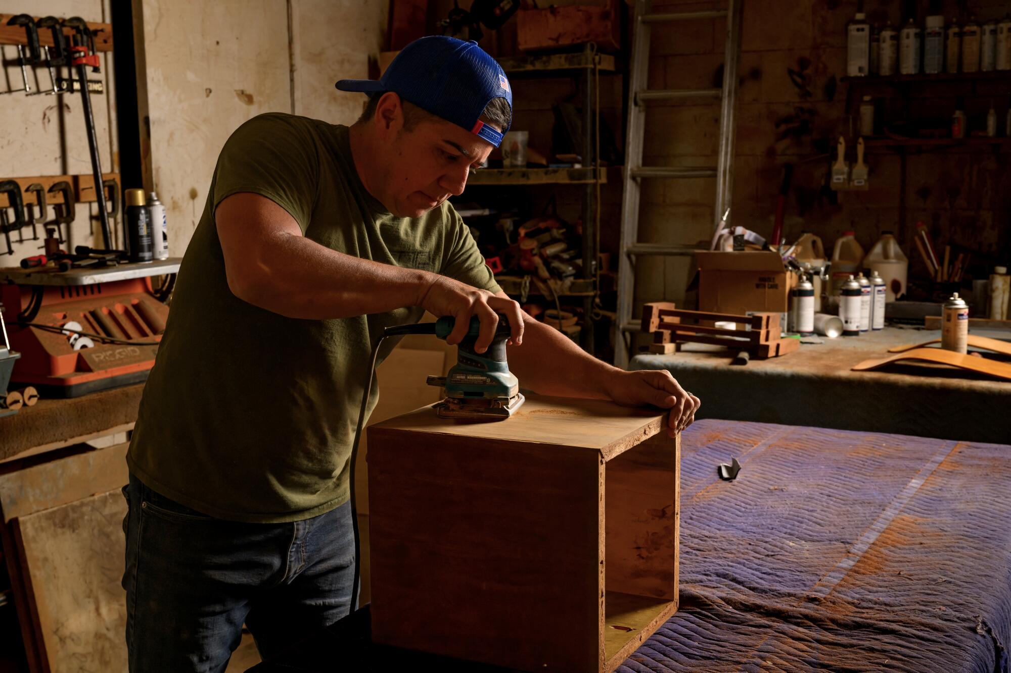 A man with a blue hat and a green T-shirt sands a wood.