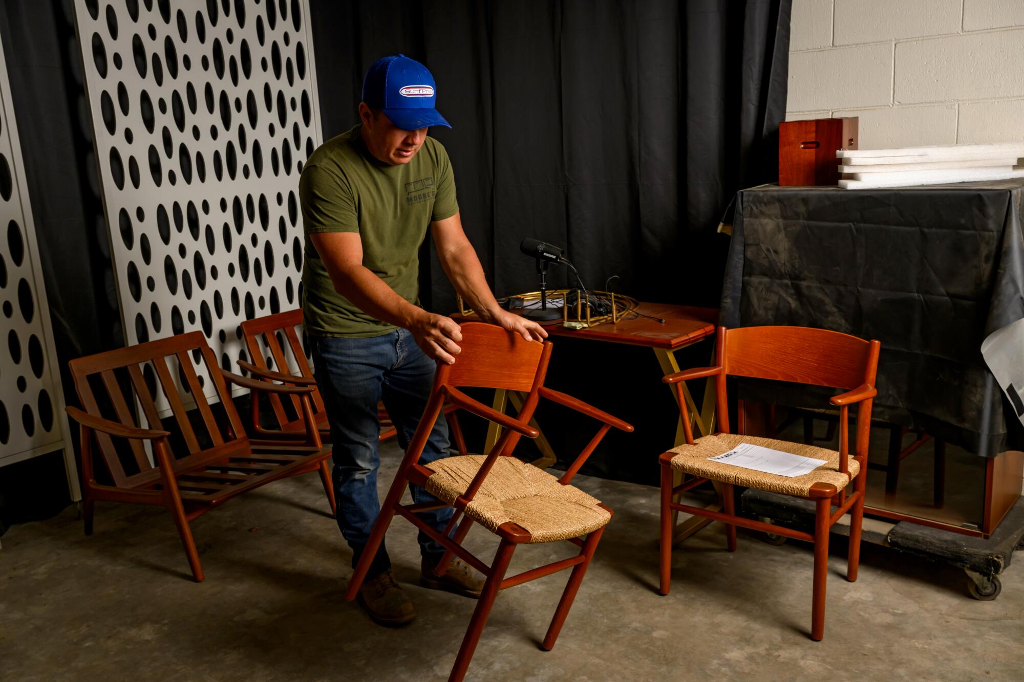 A man wearing a blue hat and green shirt hold a wooden chair in his hands.