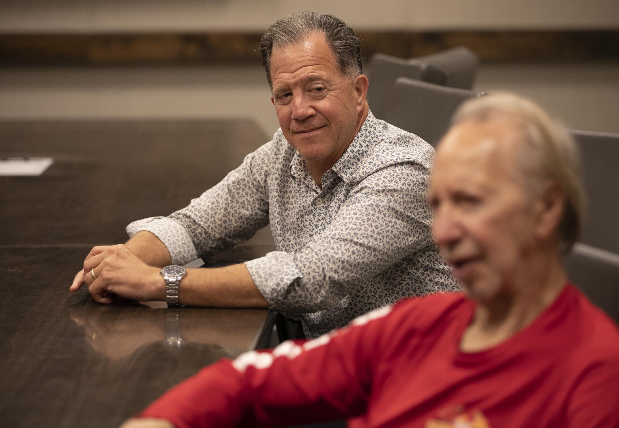 An older man and his adult son sit at a table indoors as they discuss the motivation behind his stair climbing career.