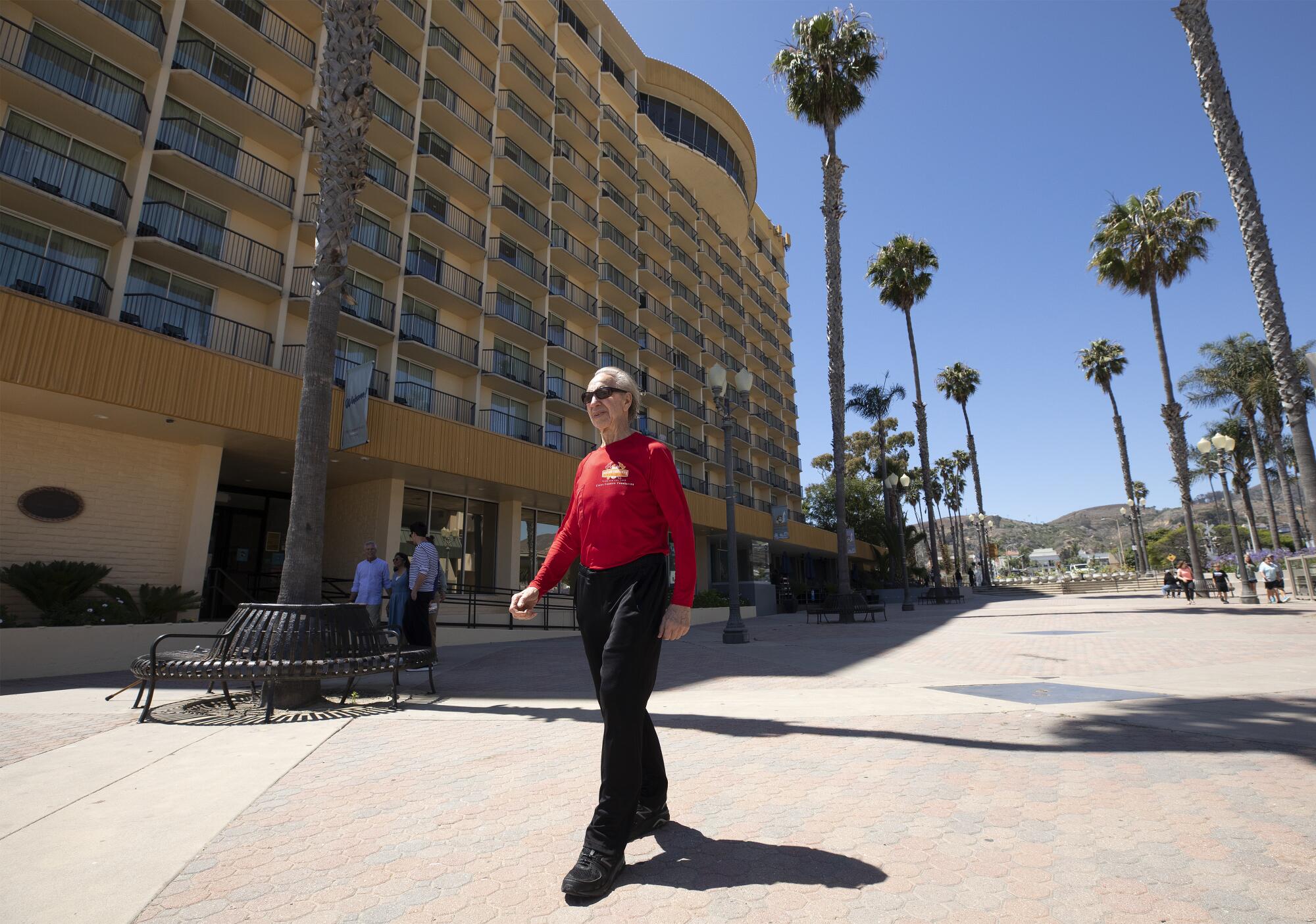 An older man walks beside a hotel on a sunny day with palm trees in the background. 