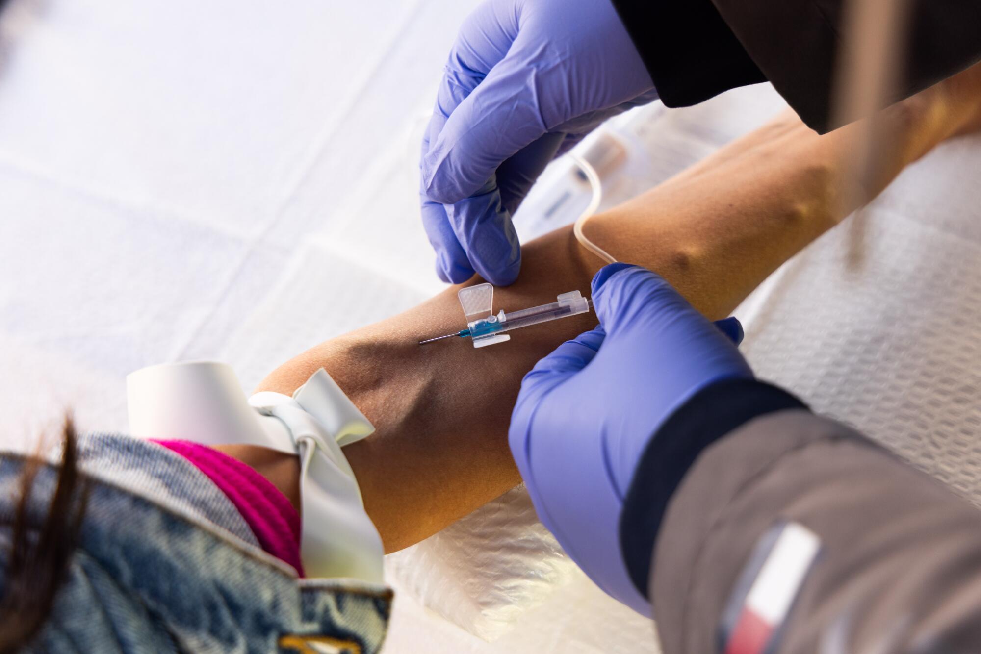 Two hands in blue gloves hold a tube on the upper arm of another person.