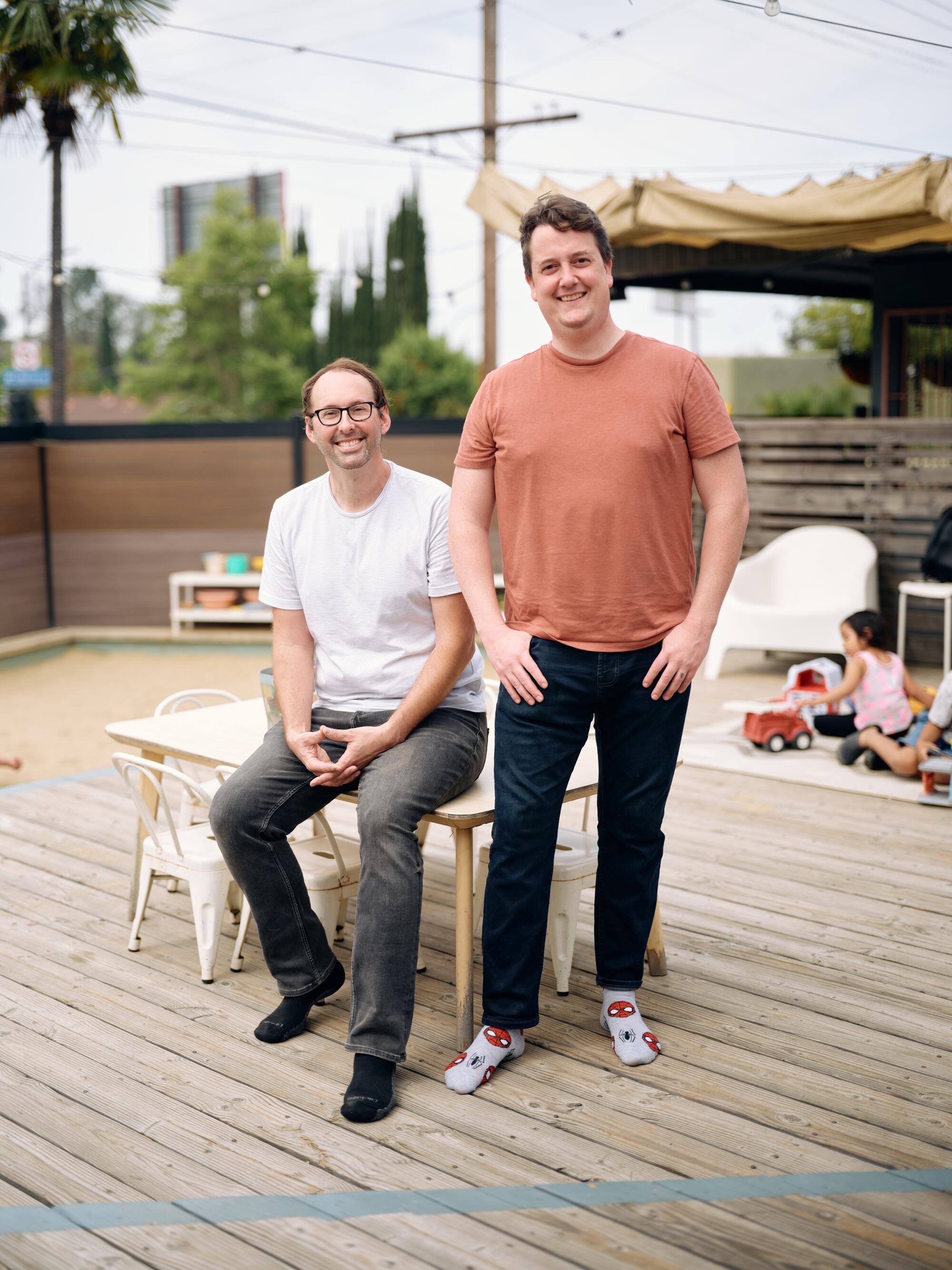 A man sits in a chair on a deck with another standing beside him. 
