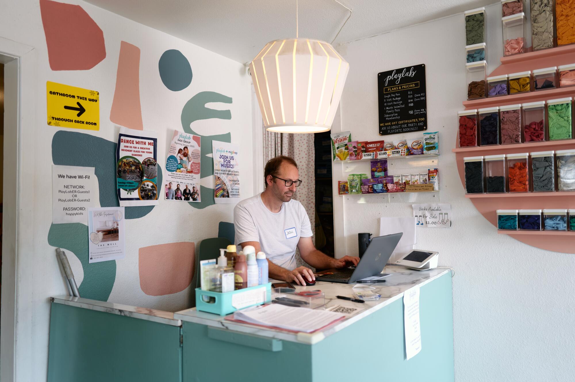 A man sits at a teal desk while working on a laptop. 