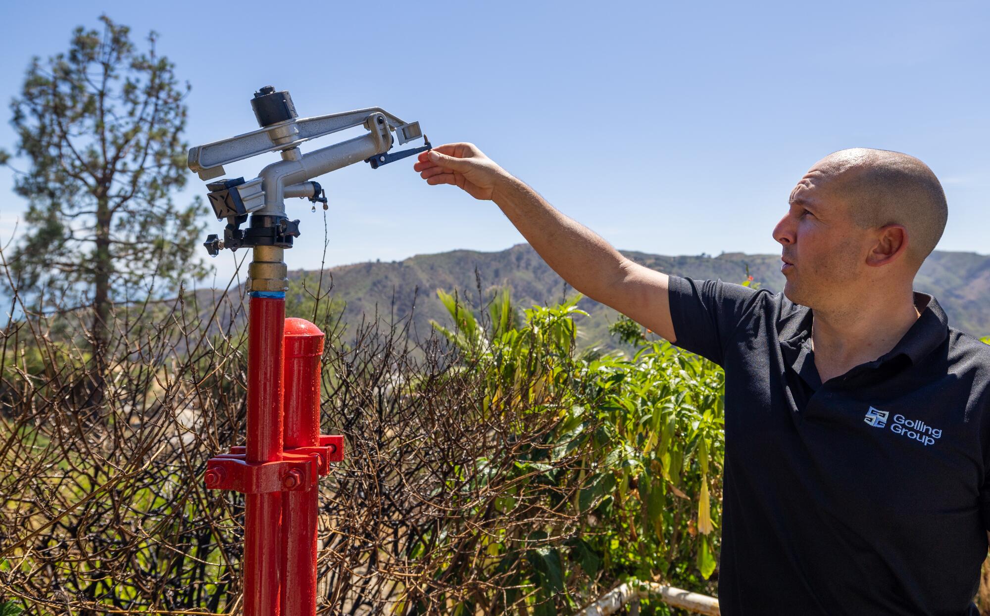 A man with a shaved head, in a dark short-sleeved shirt, adjusts a large spray nozzle on a red stand