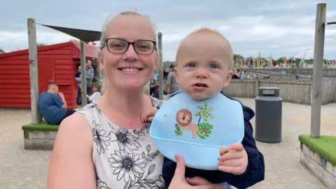Michelle Birkenhead looks straight at the camera and smiles while holding a baby wearing a blue bib with a picture of a lion on it. She is smiling, wearing glasses and a sleeveless white top with black flowers on it. She is outside and the sky behind her is overcast. 