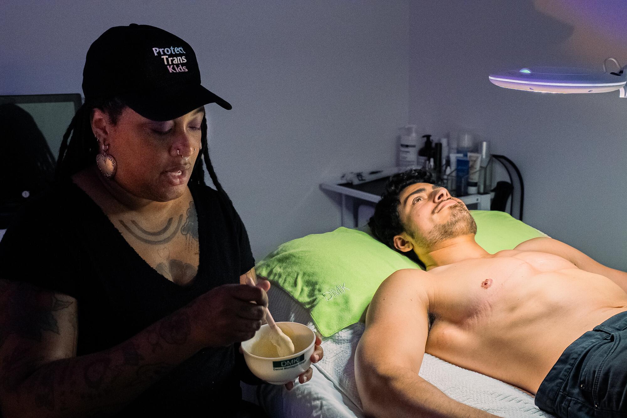 A person holds a bowl containing a skin treatment while another person lies on a table.