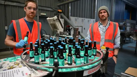 Getty Images Martin Dickie - who has short brown hair and heavy stubble - and an unshaven James Watt, who is wearing a grey beanie hat, (right) pictured packing bottles of BrewDog hand sanitizer during the Covid pandemic in April 2020. The black bottles have green labels and light blue tops. Both men are wearing orange hi-viz vests. Dickie is also wearing clear safety glasses.