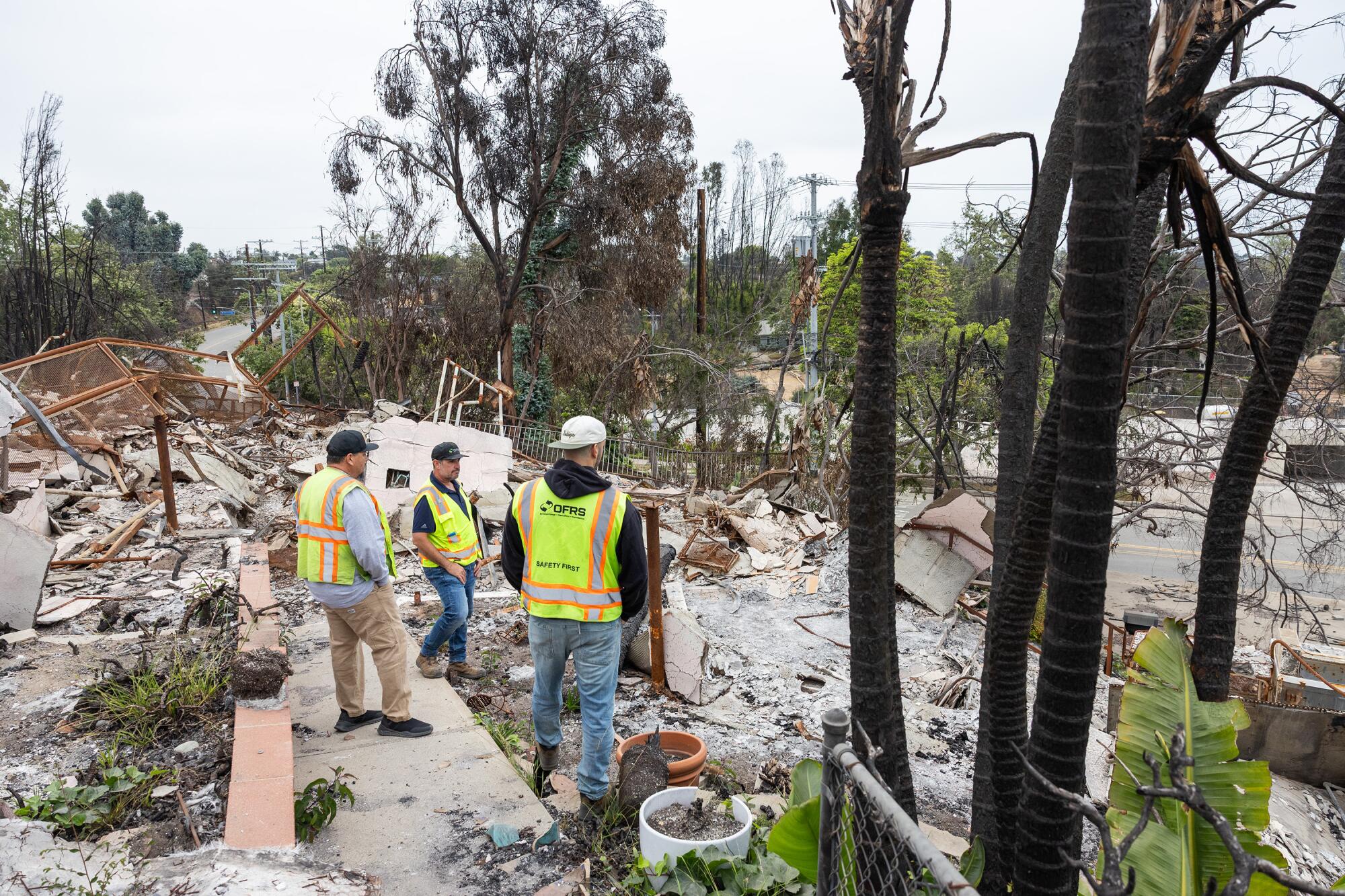 Three people wearing reflective work vests stand on a burned property