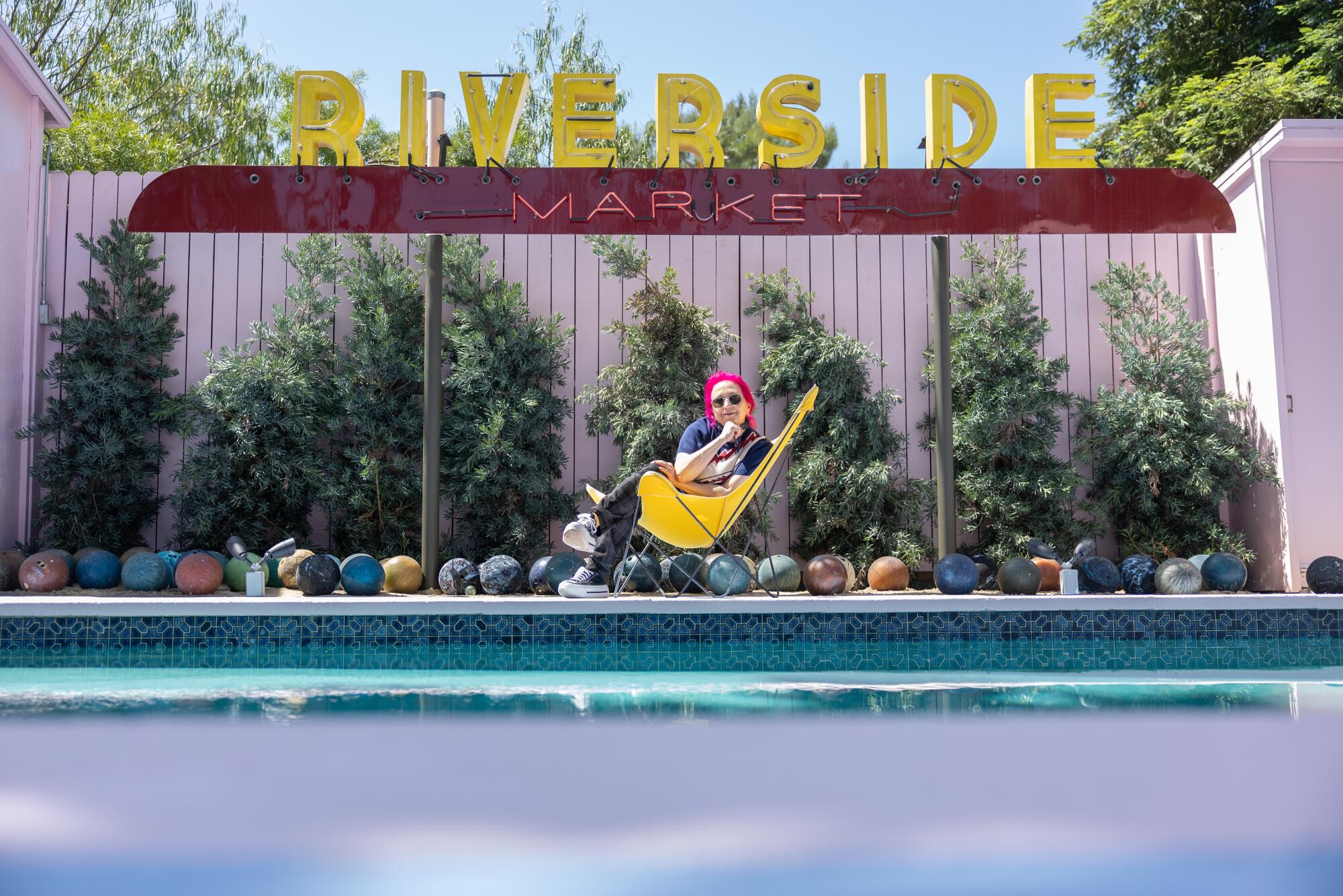A woman with pink hair lounges by a pool lined with bowling balls under a sign that says Riverside Market.