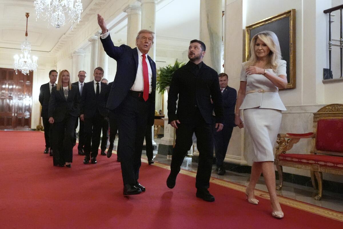 A dark-bearded man, in dark suit, walks with another man, in suit and red tie, and a woman in a white suit