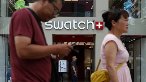 Reuters People walk past a store of Swiss watchmaker Swatch, in Beijing, China. A woman and a man can be seen in the foreground. He is wearing a maroon t-shirt and she is wearing a pink top with a yellow bag. The Swatch logo can be seen shining behind them, with the entrance to the store below