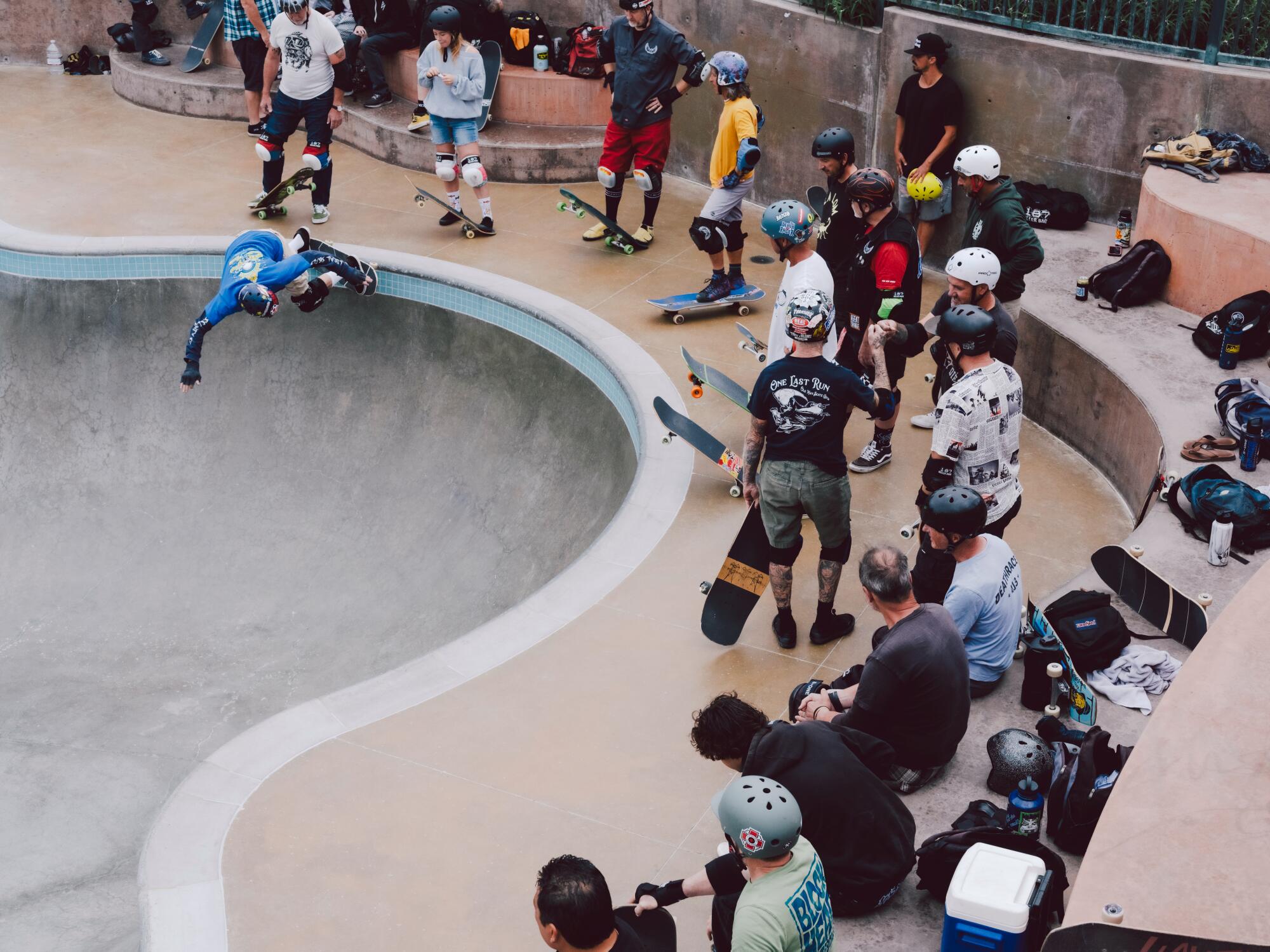 Skateboarders mingle at a skate park as one of them drops into the pool bowl.