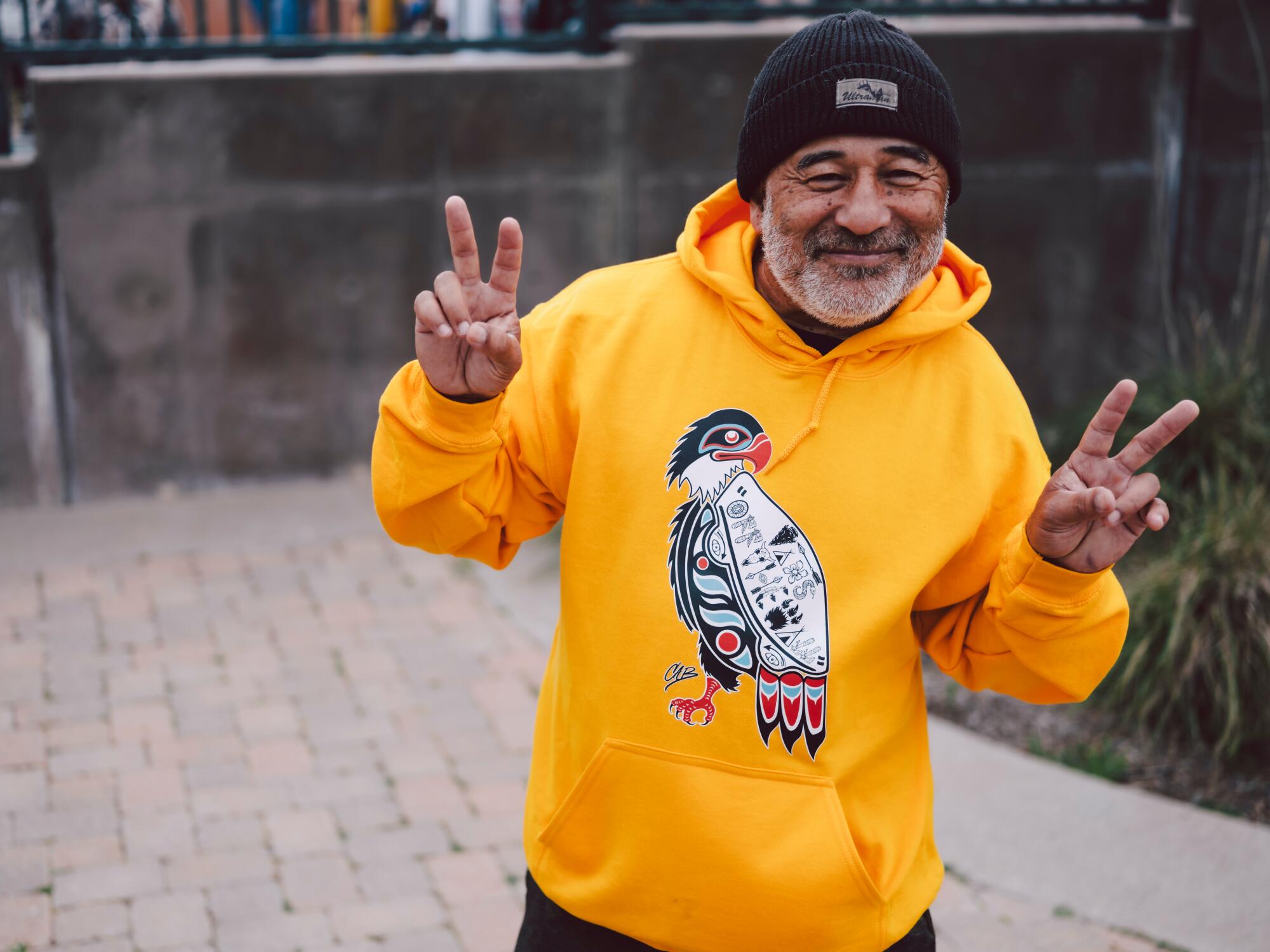 Skateboarder, Steve Caballero, poses in a yellow sweatshirt while making peace signs with his fingers.