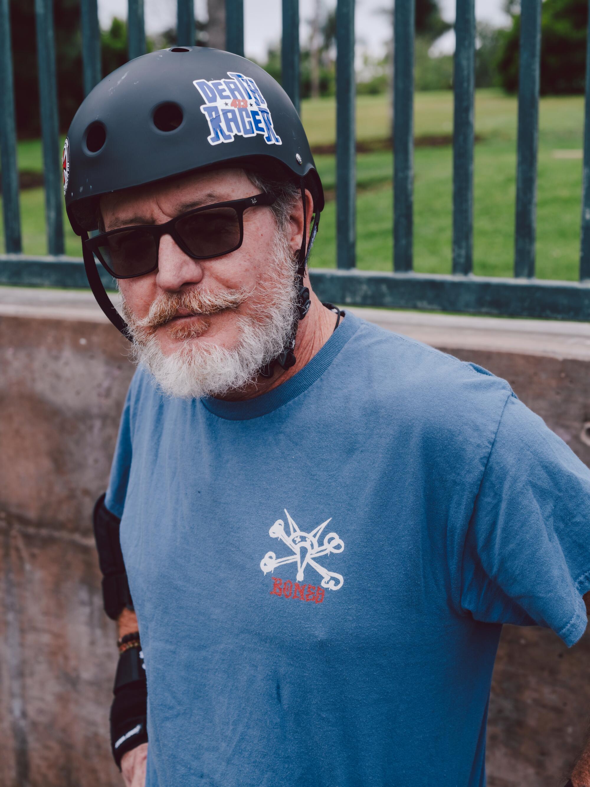 A skateboarder with a gray beard poses with hands on hips and wearing a black helmet.