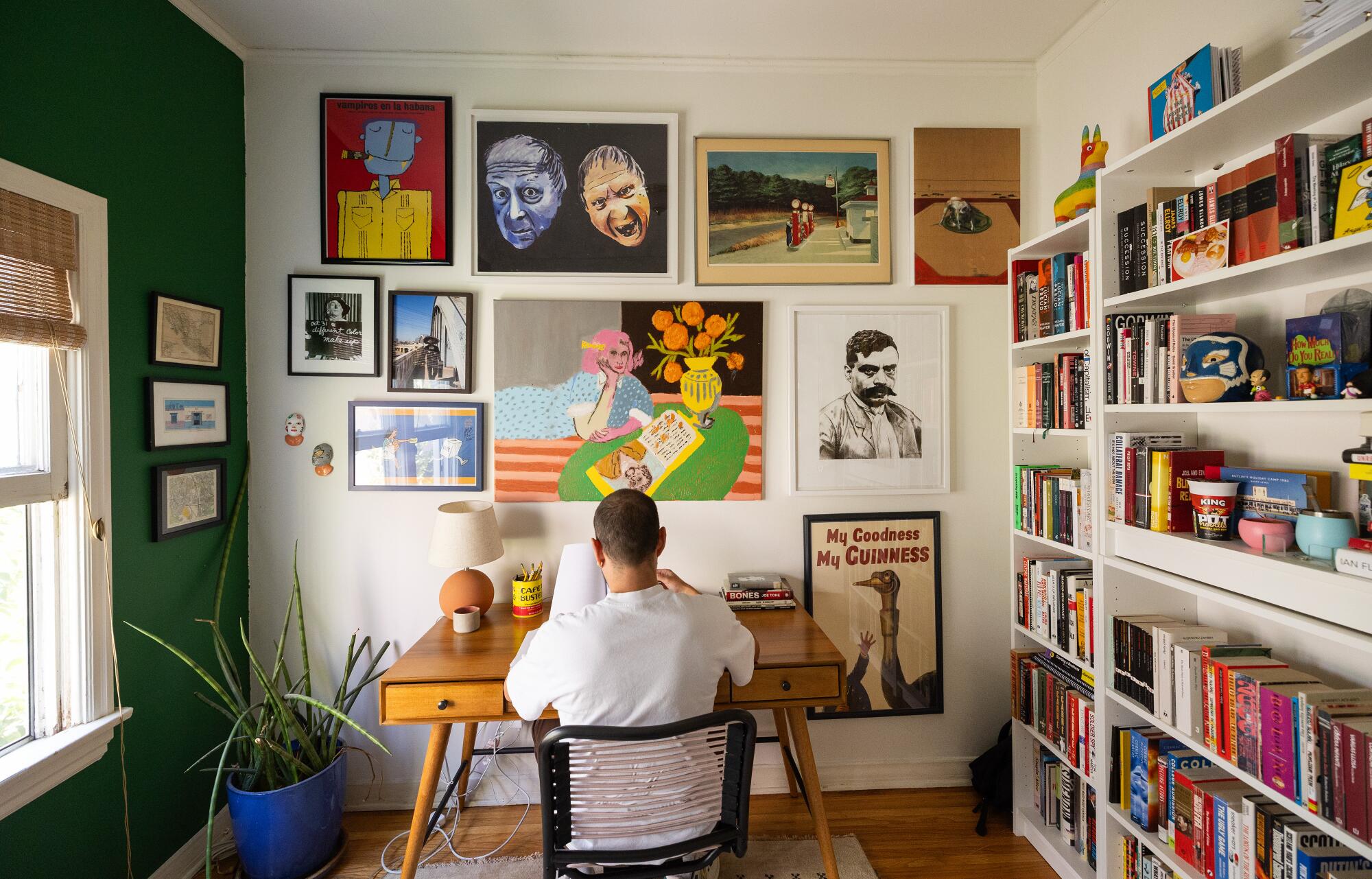 A man sits at his desk in an art-filled bedroom.