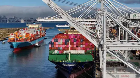 Getty Images Two ships with multi-coloured containers at the Port of Oakland, California. One of the ships is tied to the dock while the other is being escorted by two pilot boat.