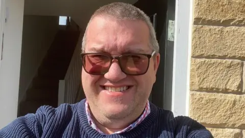 Chris Williamson Chris has short cropped grey hair and is wearing sunglasses on his face and a blue jumper over a checked shirt. He is standing in front of a house while taking this selfie.