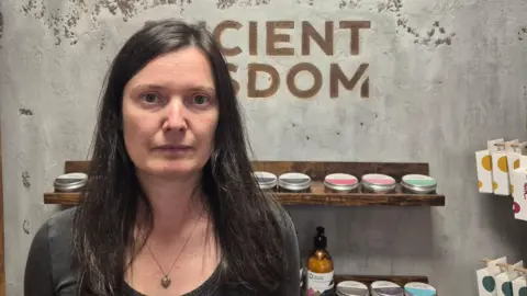 Katerina Buchy Katerina Buchy, a woman with long dark hair, wearing a dark grey top and a necklace, looks at the camera. The words "Ancient Wisdom" are written on the wall behind her in block capitals, and below that there is a shelf with cosmetic products on it.