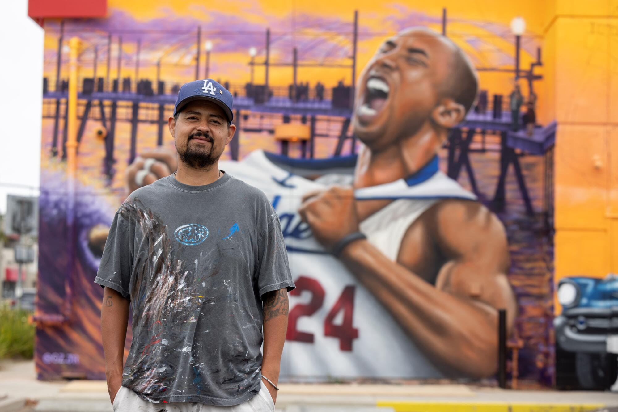 A man in a Dodgers cap and faded black T-shirt stands with his hands in his pockets in front of a Kobe Bryant mural