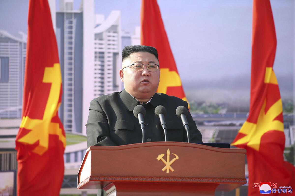 A man with dark hair, in glasses and a dark jacket, speaks at a lectern, flanked by red-and-yellow flags