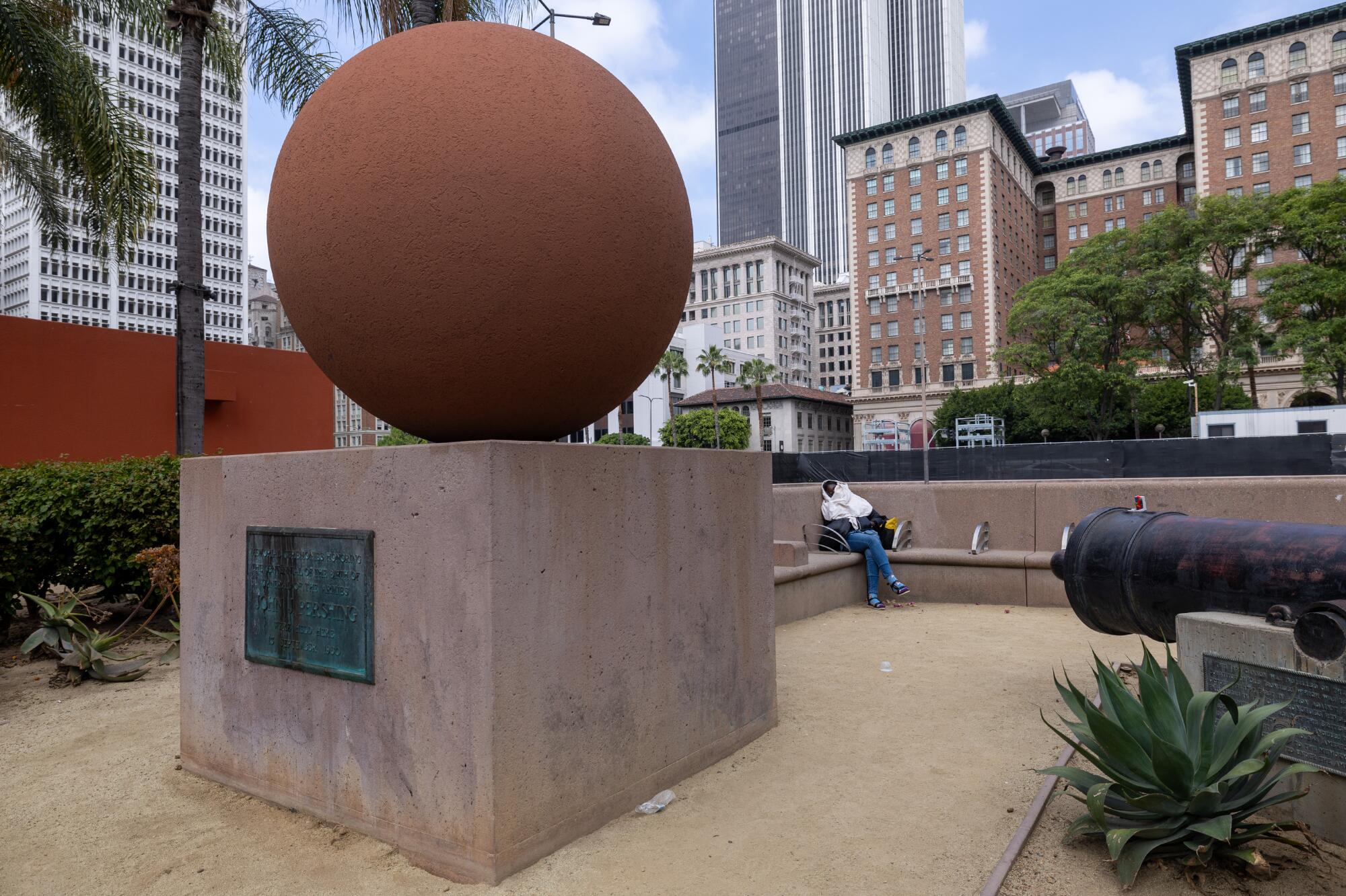 A woman sits on a bench behind a sculpture of a sphere on a square base, with high-rises as a backdrop