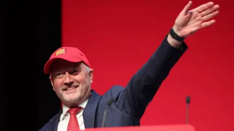 EPA Housing Secretary Steve Reed imitates US President Donald Trump's red Make America Great Again baseball caps with his own version, reading Build baby Build. He also copies Trump's navy suit, white shirt and red tie combination. He has a stubbly beard and is smiling and waving. 