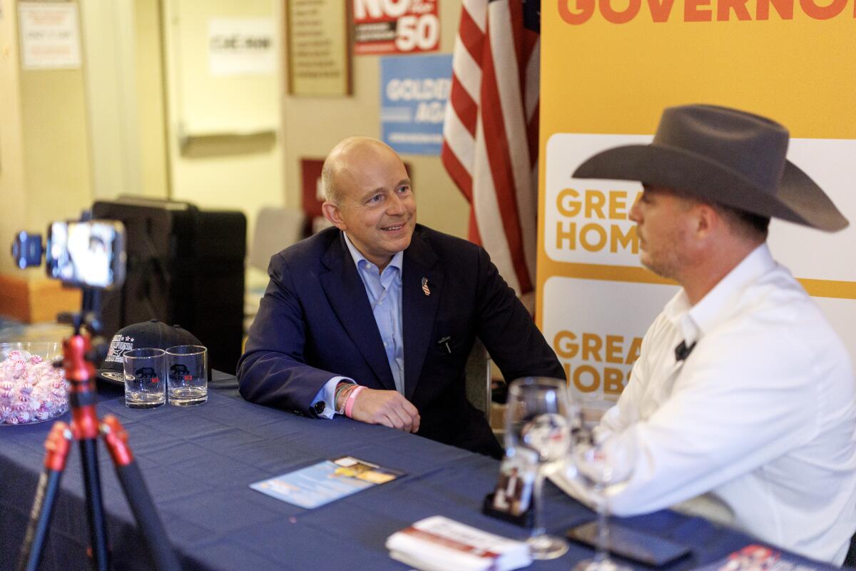 A man in a suit and a man in a cowboy hat sit next to each other at a table.