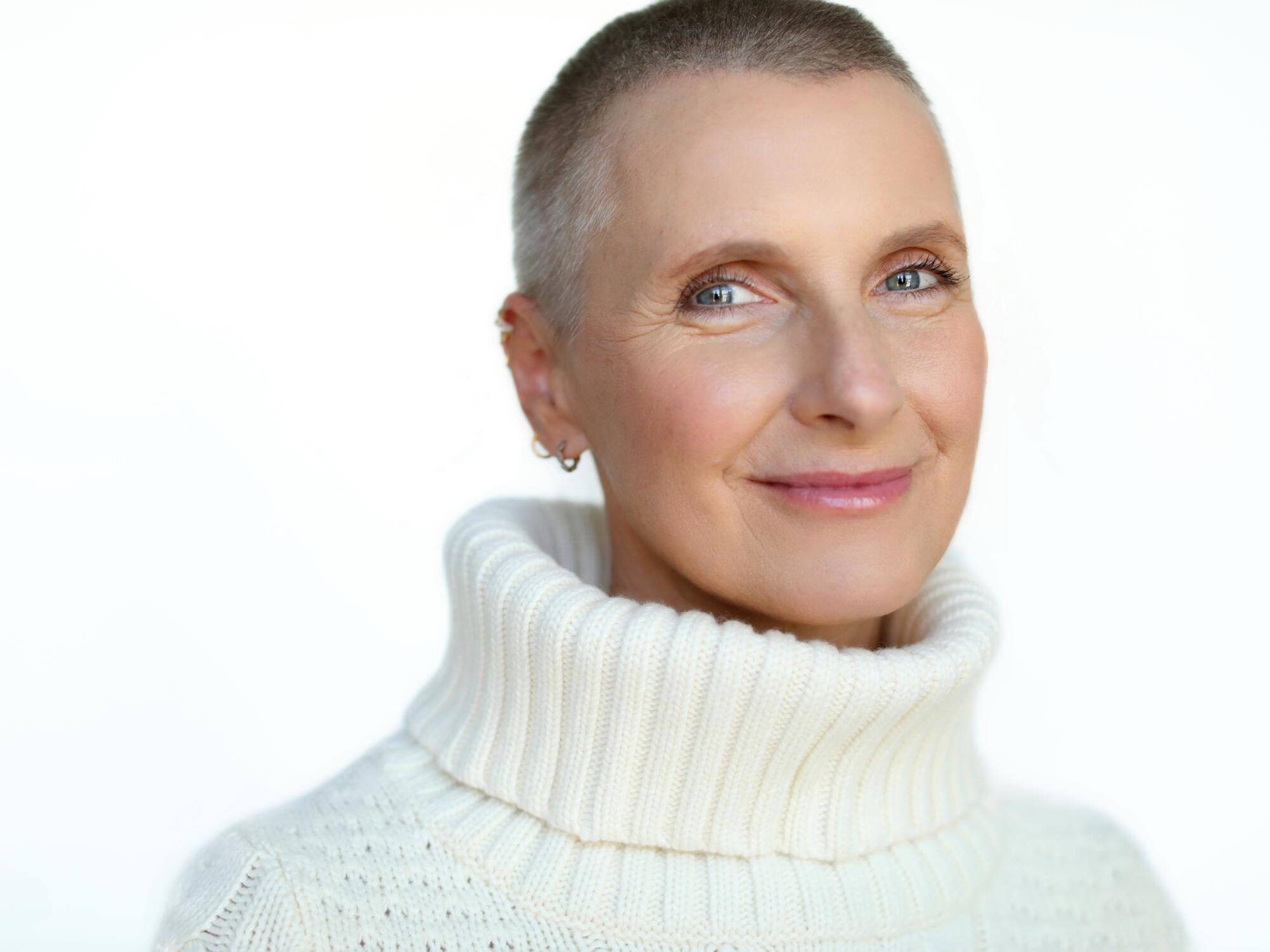 Elizabeth Gilbert, with a buzz cut and wearing a white cable-knit turtleneck sweater, smiles into the camera.