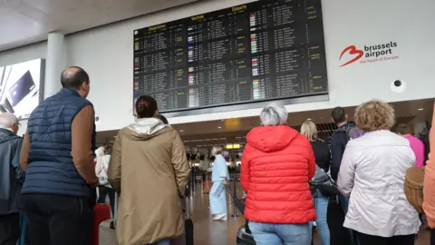 EPA A group of people leaning on wheeled suitcases look up at an airport departures board. In front of them are a series of airport check-in desks.