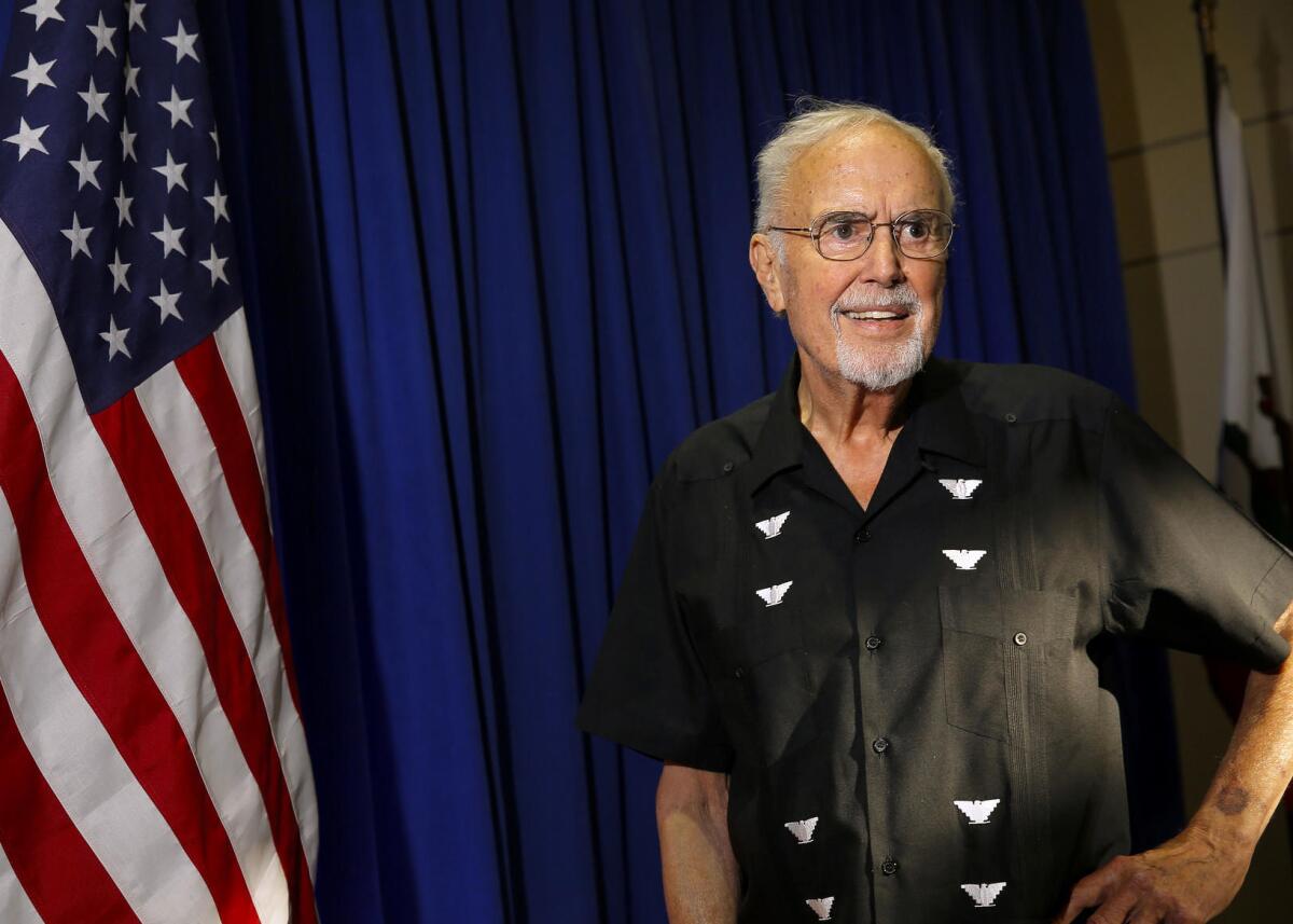 Burton wears a short-sleeved black shirt and stands near a U.S. flag.