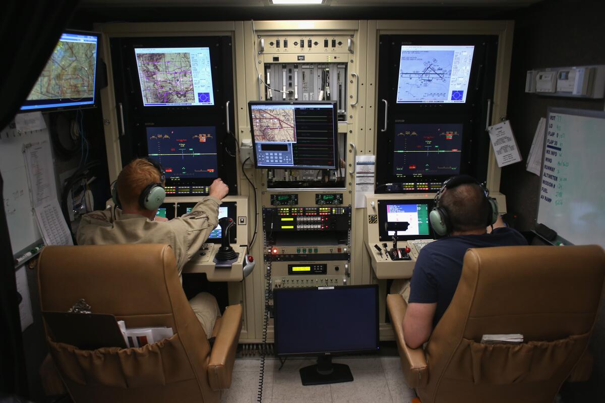 Two people in chairs look at screens and panels of buttons.