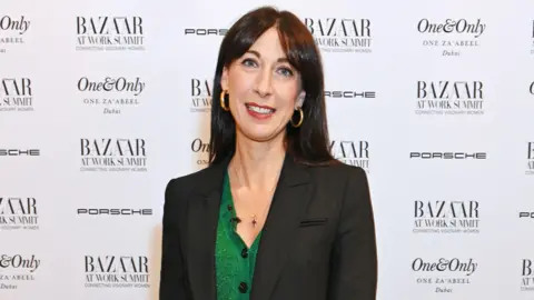 Dave Bennett/Getty Images Samanthan Cameron wearing a green cardigan under a black blazer, at a magazine event with the promotional board behind her. She is smiling and wearing gold hoop earrings.