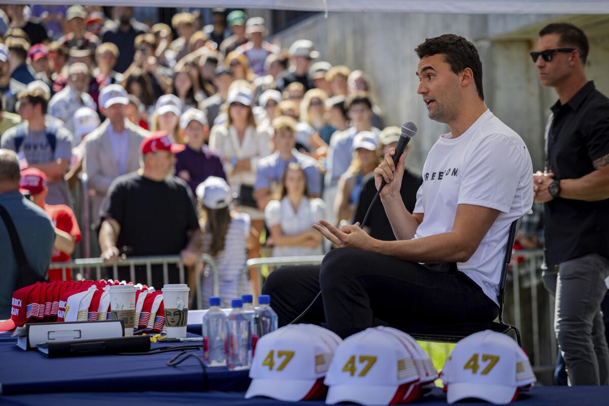 Charlie Kirk speaks to an audience, seated next to stacks of hats reading "47."