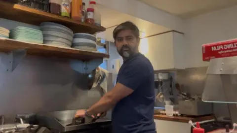 Prasanna Callaghan A middle aged man with a greying beard stands in a cafe kitchen. He is staring into the camera. There are many plates and condiment bottles on shelves in front of him. 