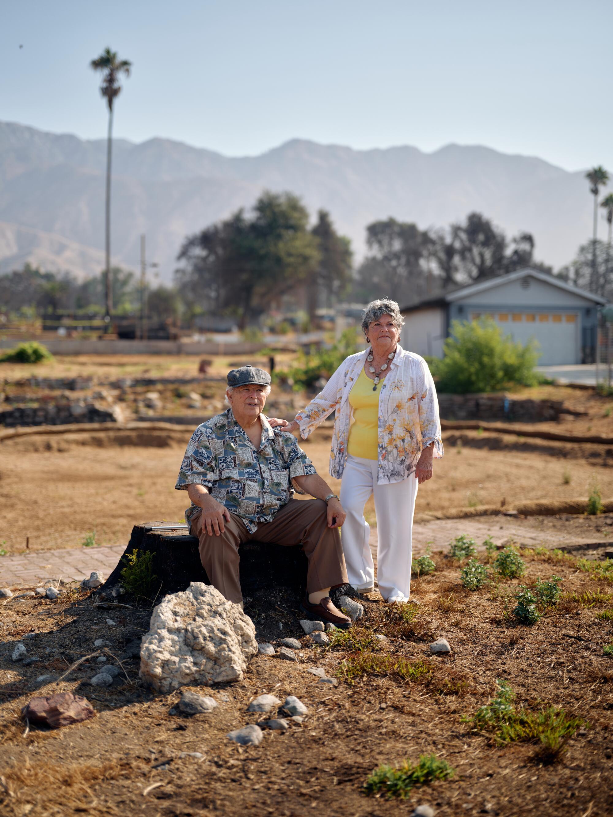 A man sits on a broad tree stump, his wife stands beside him, in front of a barren area where their home once stood.