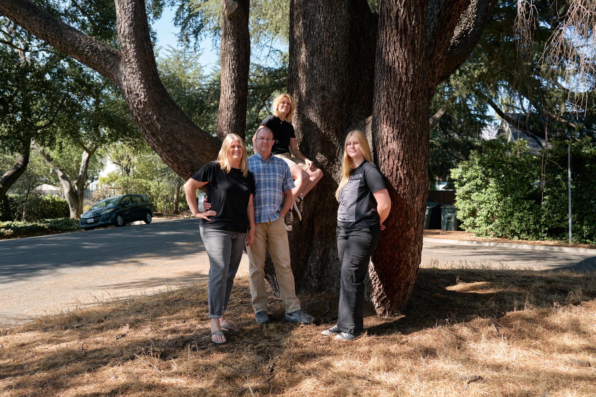 A blond woman, her husband and their two blond young adult daughters pose against a deodar cedar tree.